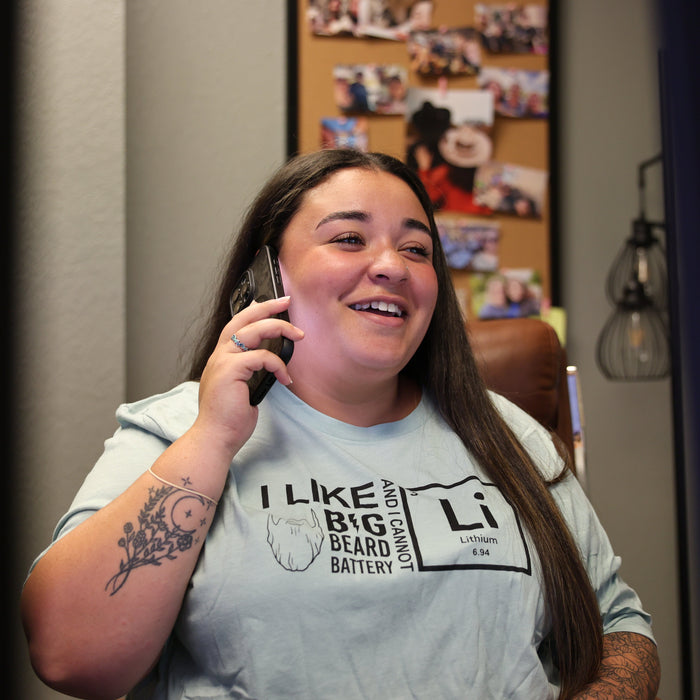 A woman with long dark hair smiling while talking on a mobile phone, wearing a light blue graphic t-shirt with a black printed design that reads 'I LIKE BIG BEARD BATTERY' and a periodic table-style 'Li' logo.